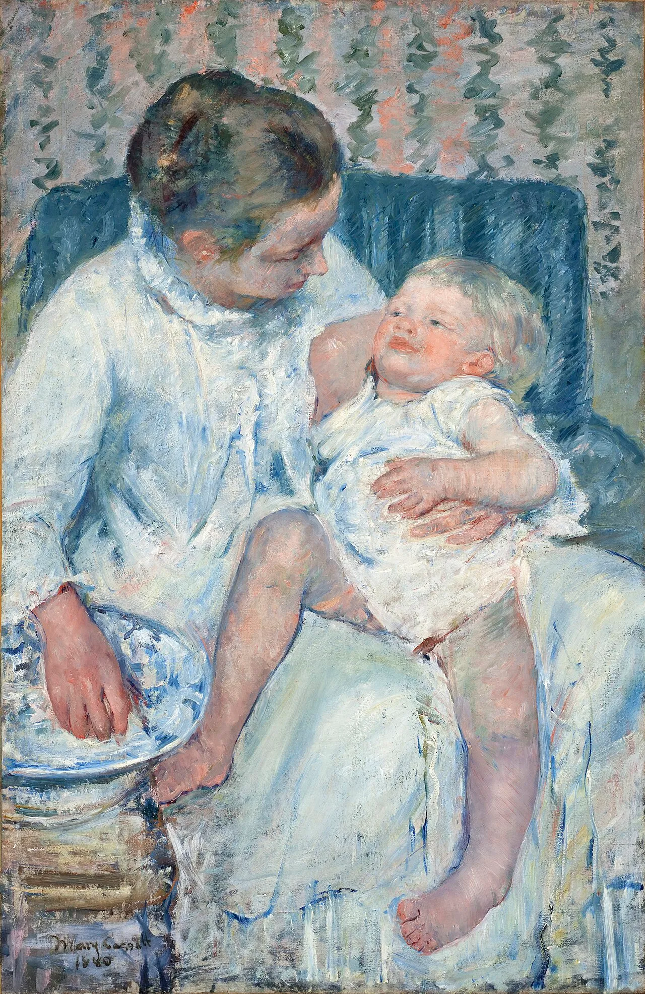 A mother in a white gown holds a smiling baby on her lap beside a washing bowl