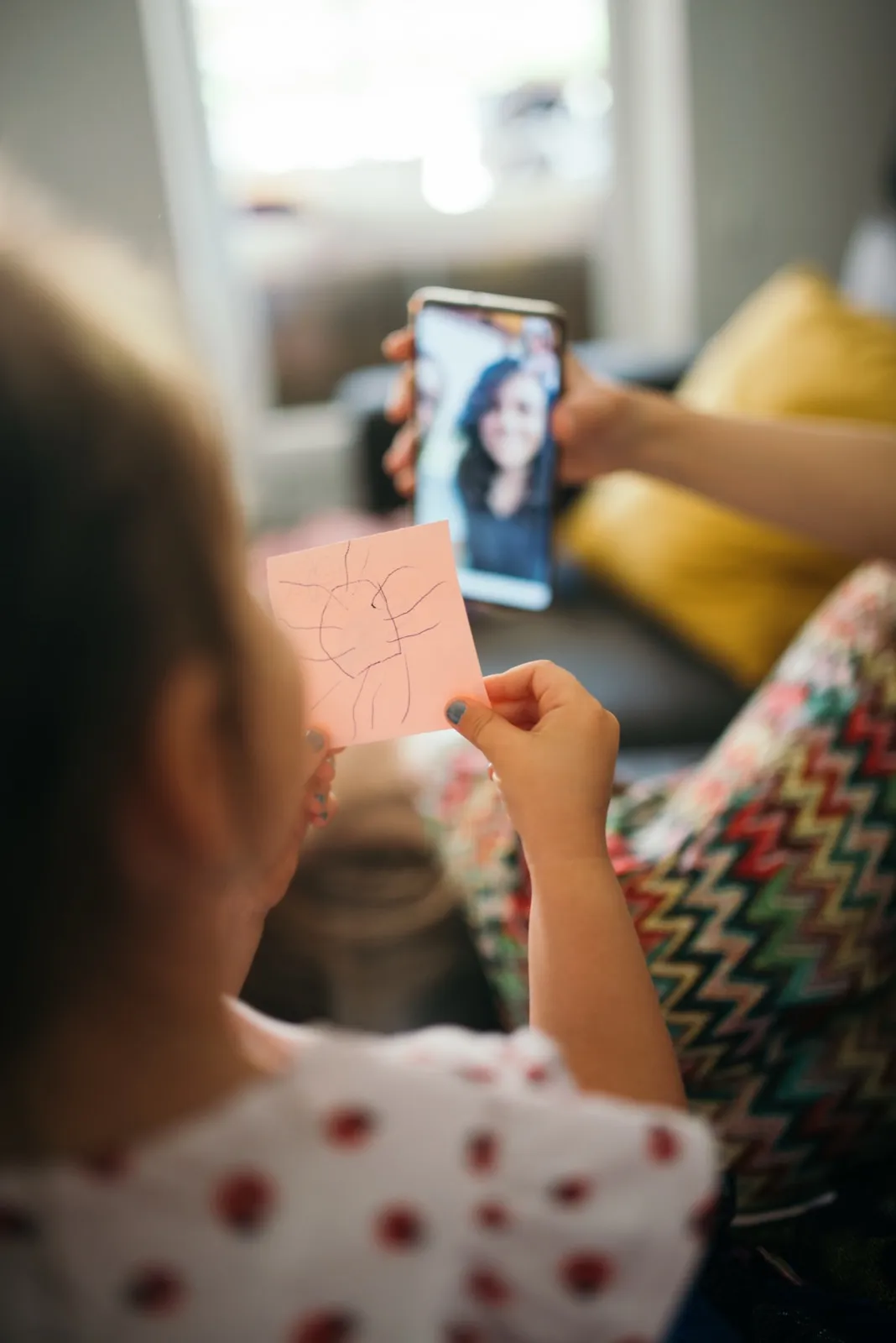 A small child holds up a drawing to a smiling woman on a smartphone video call