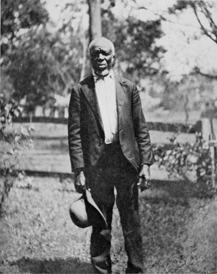 A black and white photograph of Cudjo Lewis, an elderly man in a dark suit and white shirt, standing in a garden holding a hat