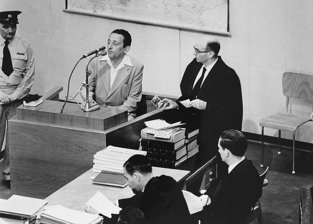 Prosecutor Gideon Hausner standing beside the witness box, holding up a photograph as the witness Henryk Ross answers at the microphone