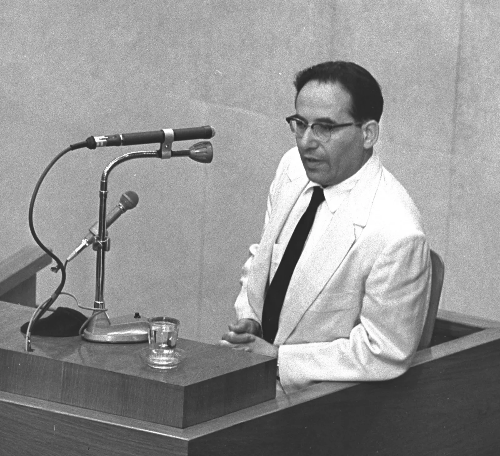 Yehiel De-Nur, a survivor of Auschwitz, in a light suit and dark tie, testifying at a microphone in the witness box at the Eichmann trial in Jerusalem