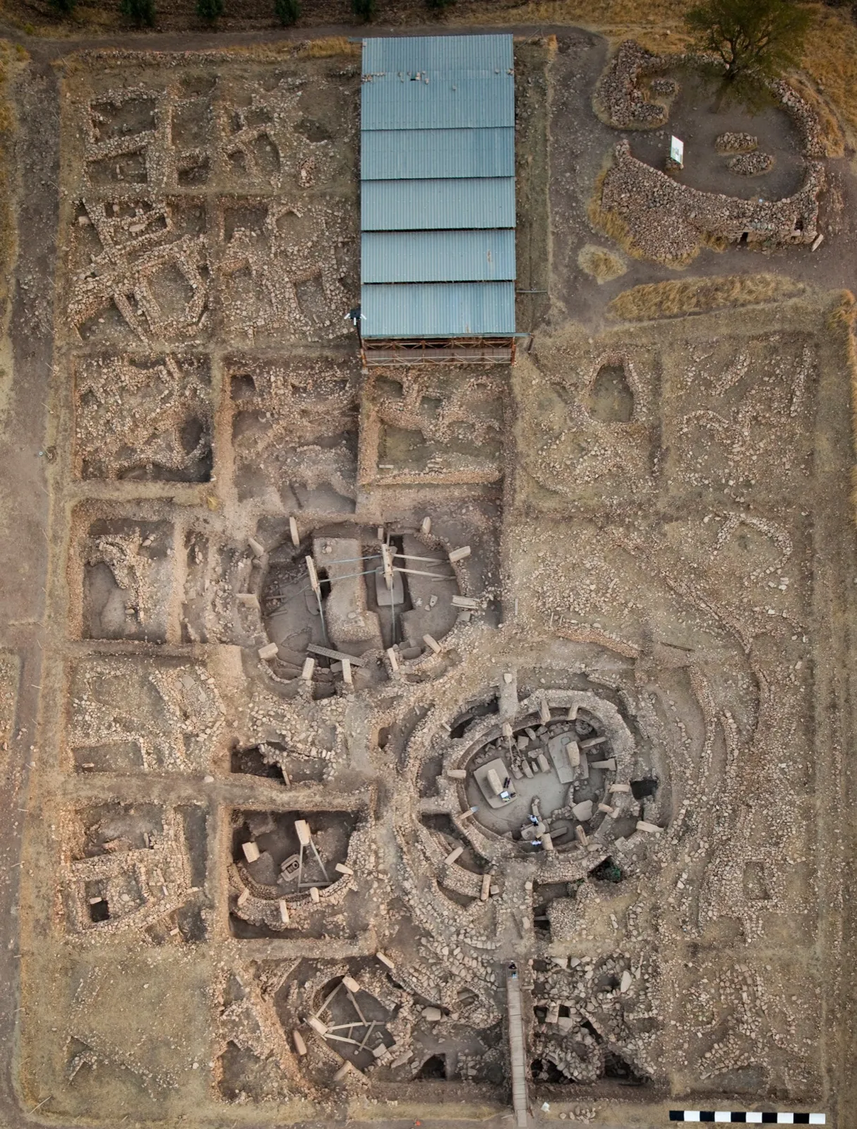An aerial photograph of the main excavation area at Göbekli Tepe, showing several circular stone enclosures ringed by T-shaped pillars, surrounded by rectangular trenches and a protective roof