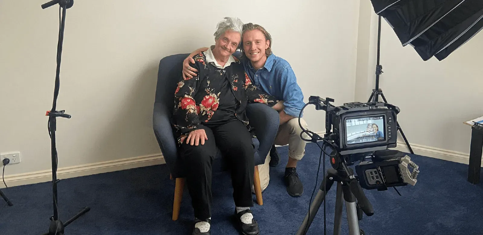 Daniel with his arm around his grandmother after filming her life story, beside the camera and studio lights