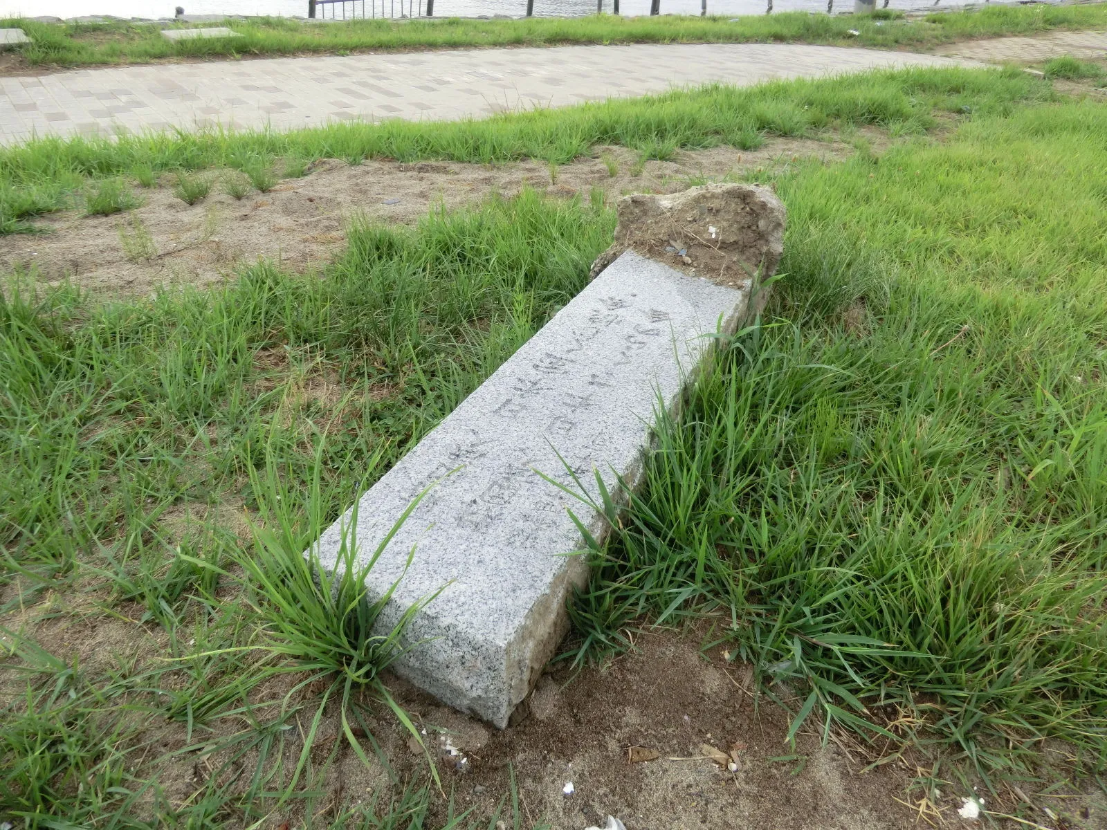 A fallen granite monument lying in grass beside a paved path, with carved Japanese characters on its side