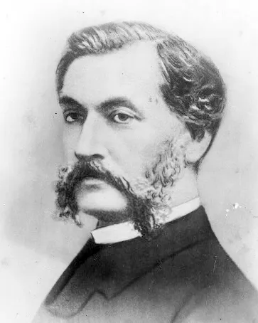 A black-and-white portrait photograph of a man with dark wavy hair, a heavy moustache and long side whiskers, wearing a dark jacket and white collar