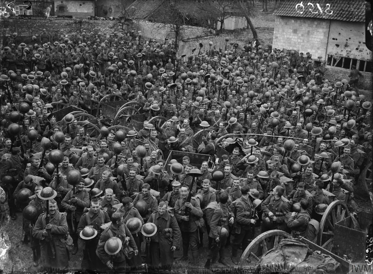 Several hundred British soldiers in a farmyard, smiling and holding their steel helmets up on the ends of their rifles