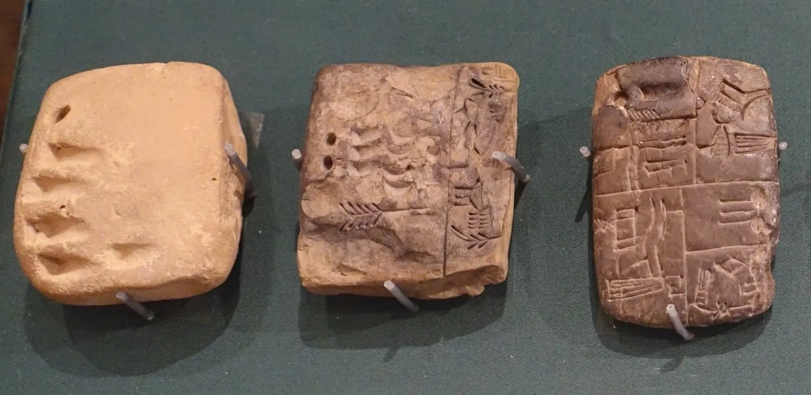 Three small clay tablets in a museum case: one with only pressed number marks, one with numbers and a few signs, and one covered in proto-cuneiform signs