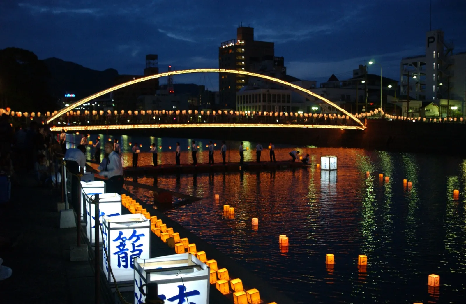 Paper lanterns floating on the Sasebo River at night during the Obon festival in Japan, with a lit arched bridge behind