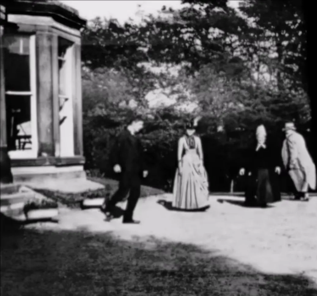 A grainy black-and-white film frame of four people in Victorian clothes walking on a sunlit lawn beside a conservatory, with trees behind them