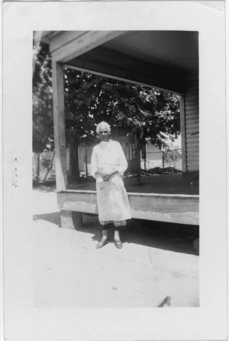 An elderly woman in a long white dress standing in front of the verandah of a timber house