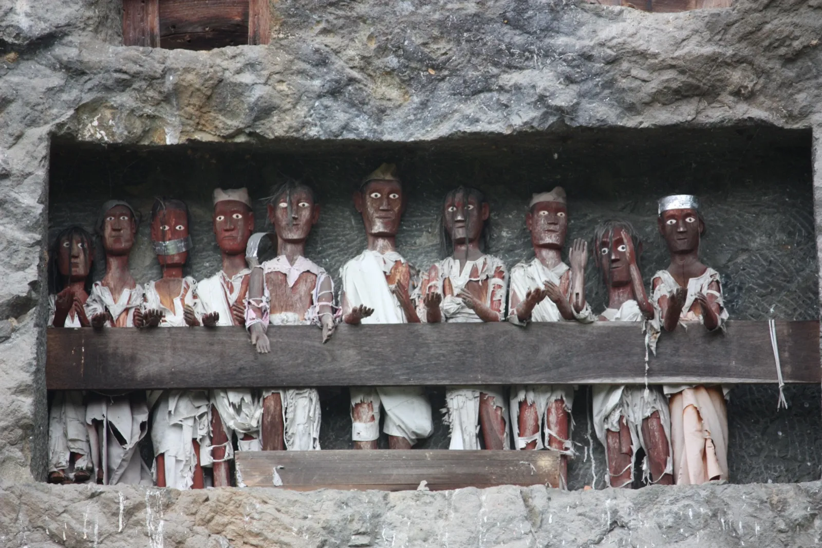 A row of carved wooden tau tau effigies of the dead, dressed in worn white cloth, standing on a balcony cut into a cliff at Lemo in Tana Toraja, Indonesia