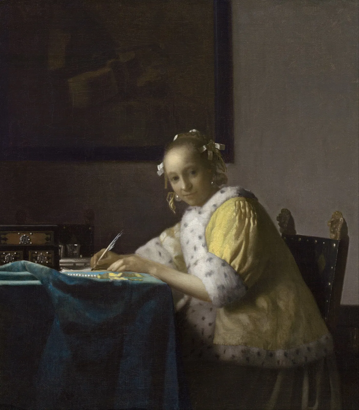 A young woman in a yellow jacket trimmed with white fur sits at a table with a blue cloth, a quill in her hand, looking up from the letter she is writing