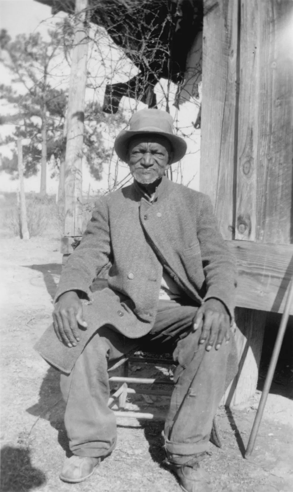 An elderly man in a herringbone coat and felt hat sitting on a chair outside a timber house