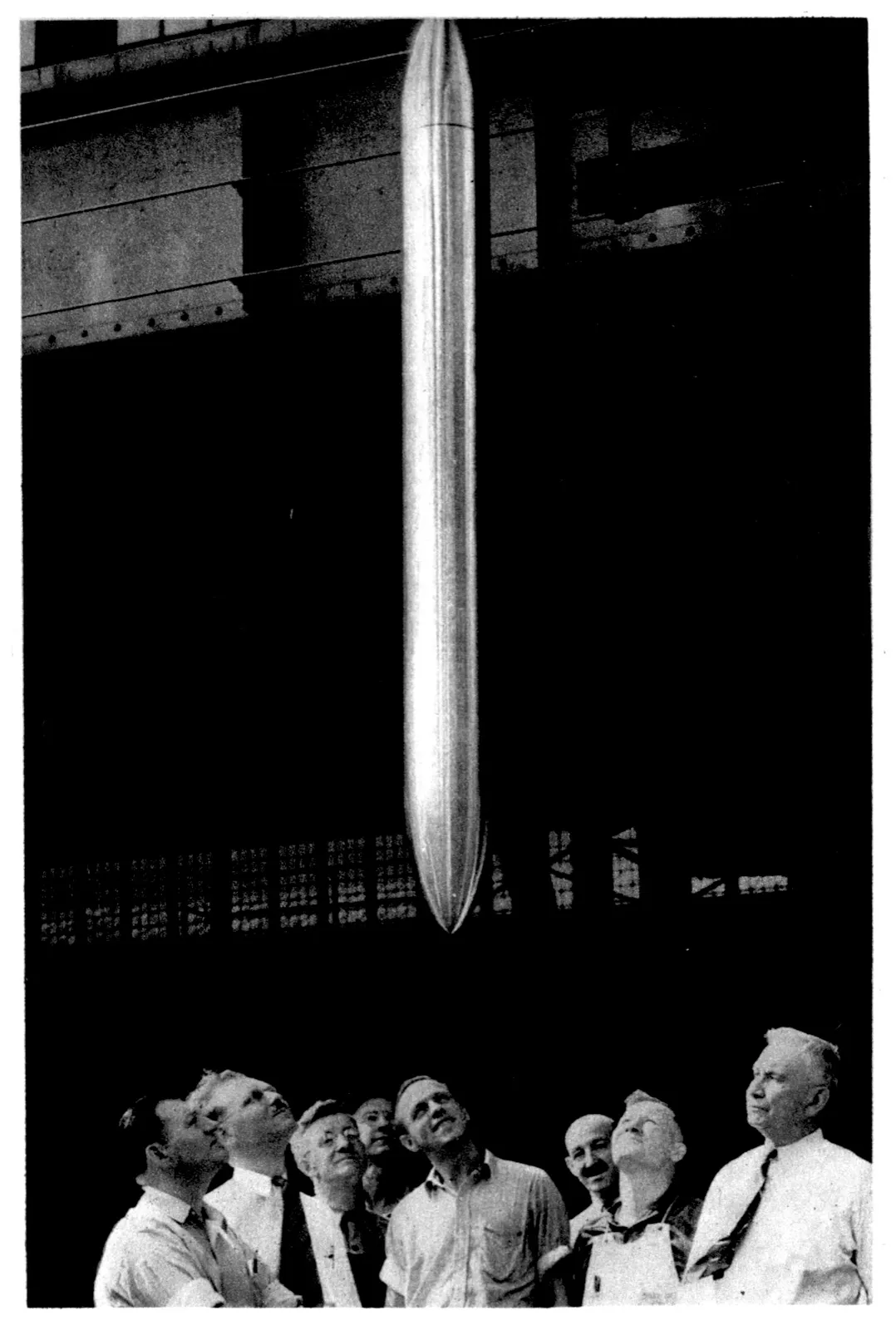 A large group of men in shirts and ties look up at a long, gleaming torpedo-shaped capsule suspended above them