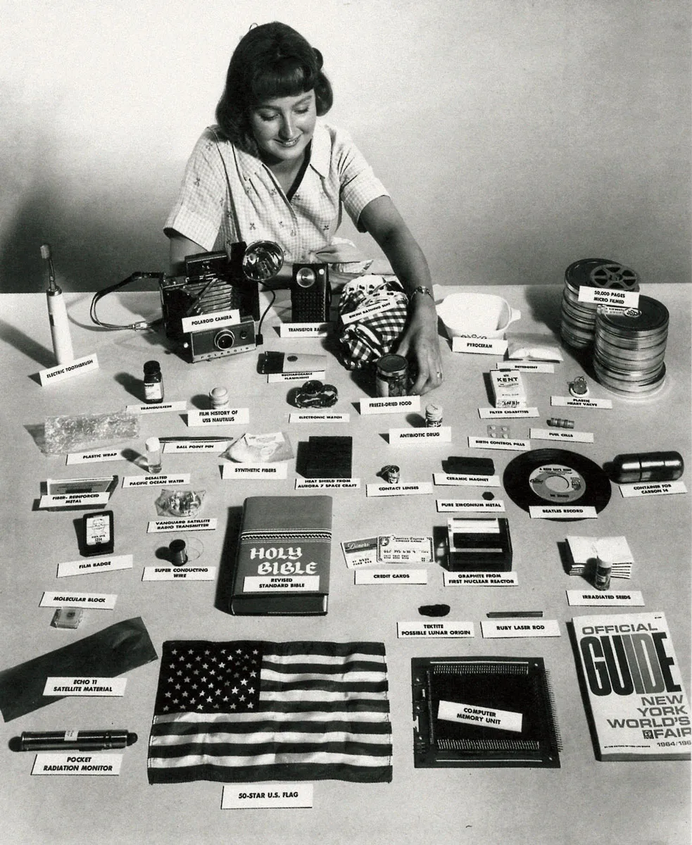 A woman arranges objects on a table for the 1965 Westinghouse time capsule, each with a label, including a Polaroid camera, a Beatles record, credit cards, a Bible, microfilm reels and a folded American flag